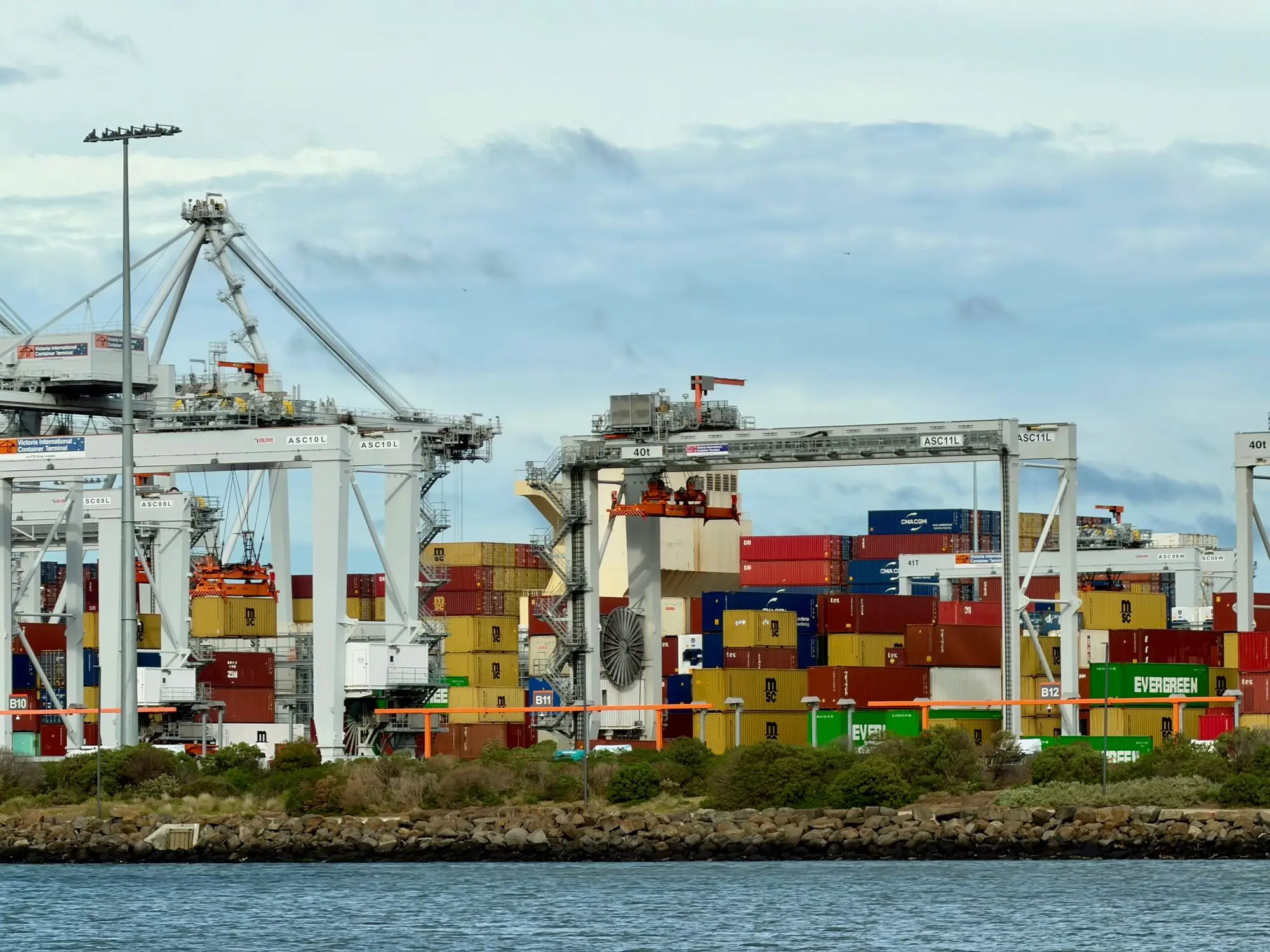 Stacked shipping containers and gantry cranes at a Port Melbourne container terminal under a cloudy sky