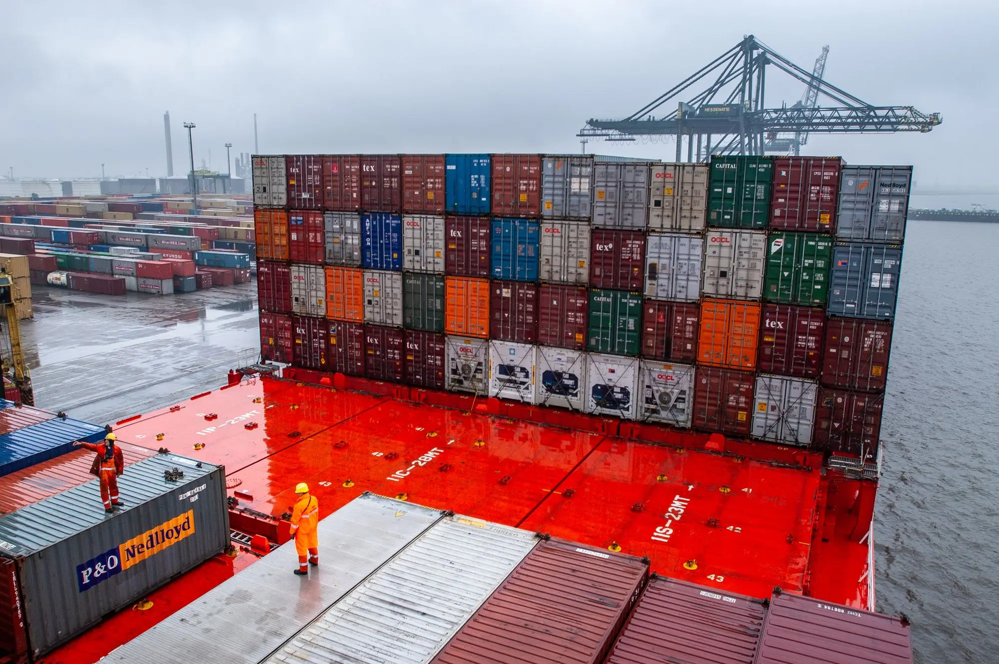 Dock workers in hi-vis on a container ship deck beside stacked containers and gantry cranes on a wet day at the terminal